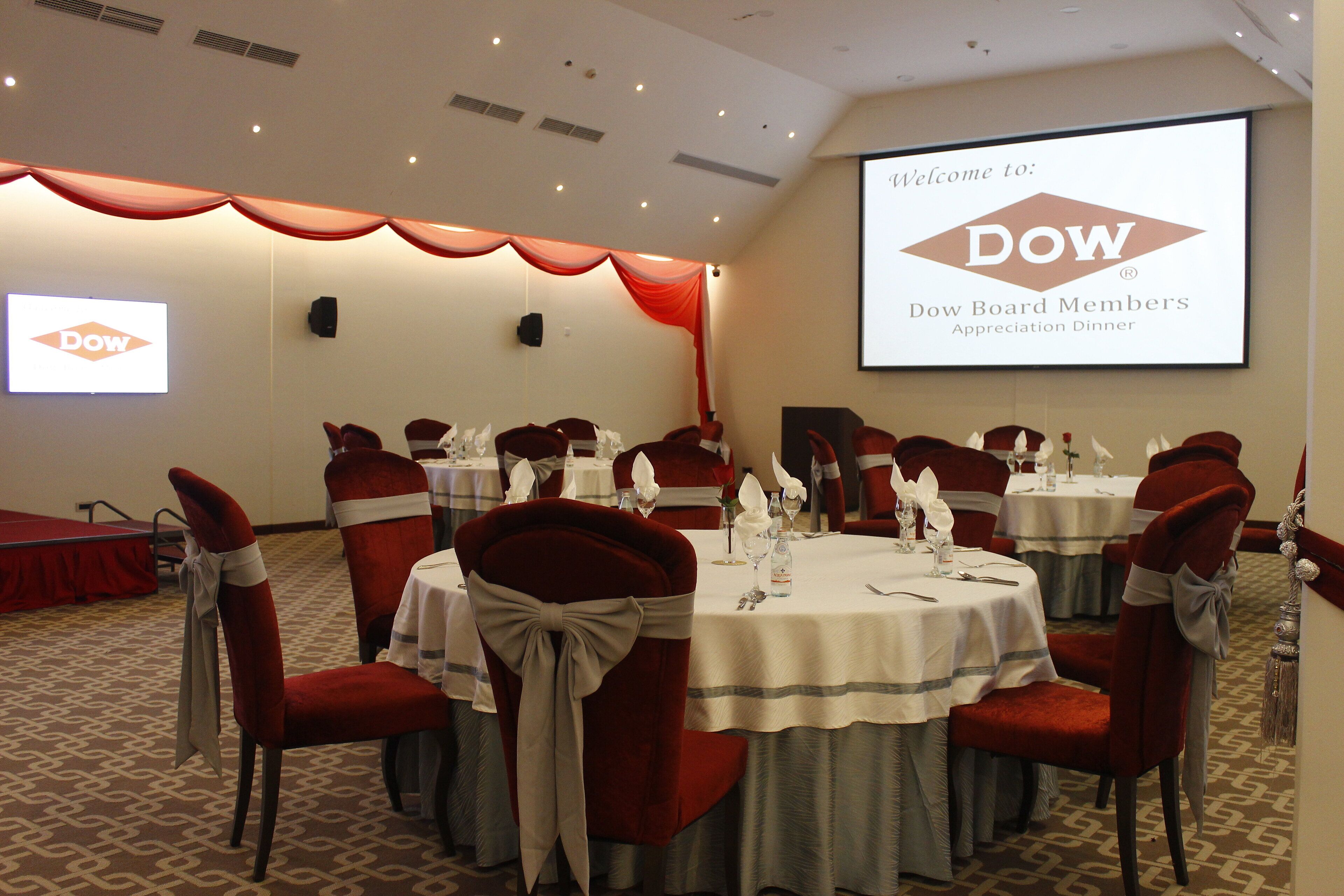 A conference room with tables set for a formal event, a screen displaying the Dow Board Members Appreciation Dinner, and a DOW logo on a wall.
