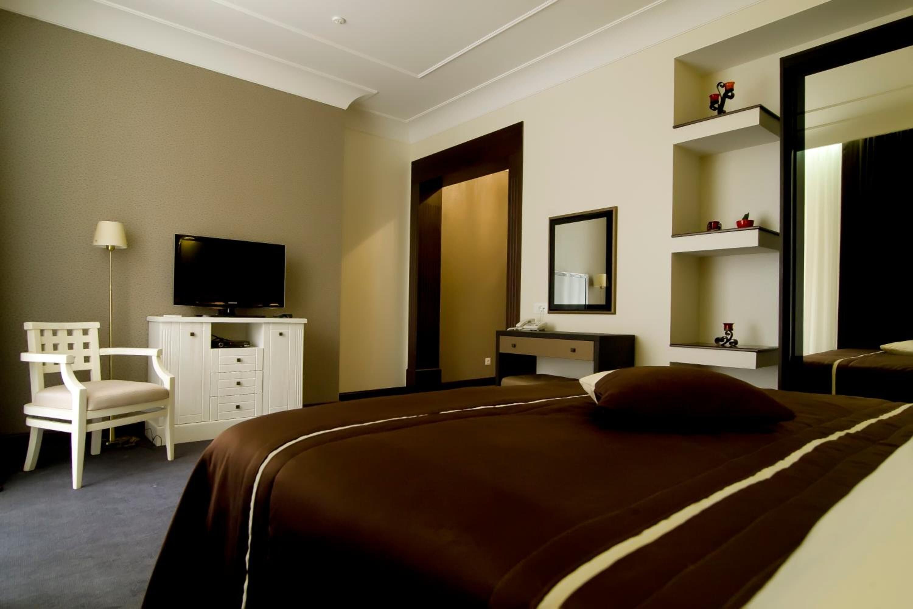 A hotel room with a bed, a desk with a chair, a television, a mirror, and a shelf with decorative items.