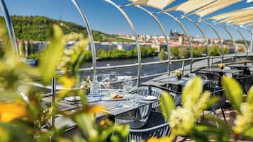 Ein Außenbereich zum Essen mit Tischen und Stühlen, Blick auf einen Fluss und einem überdachten Gang.