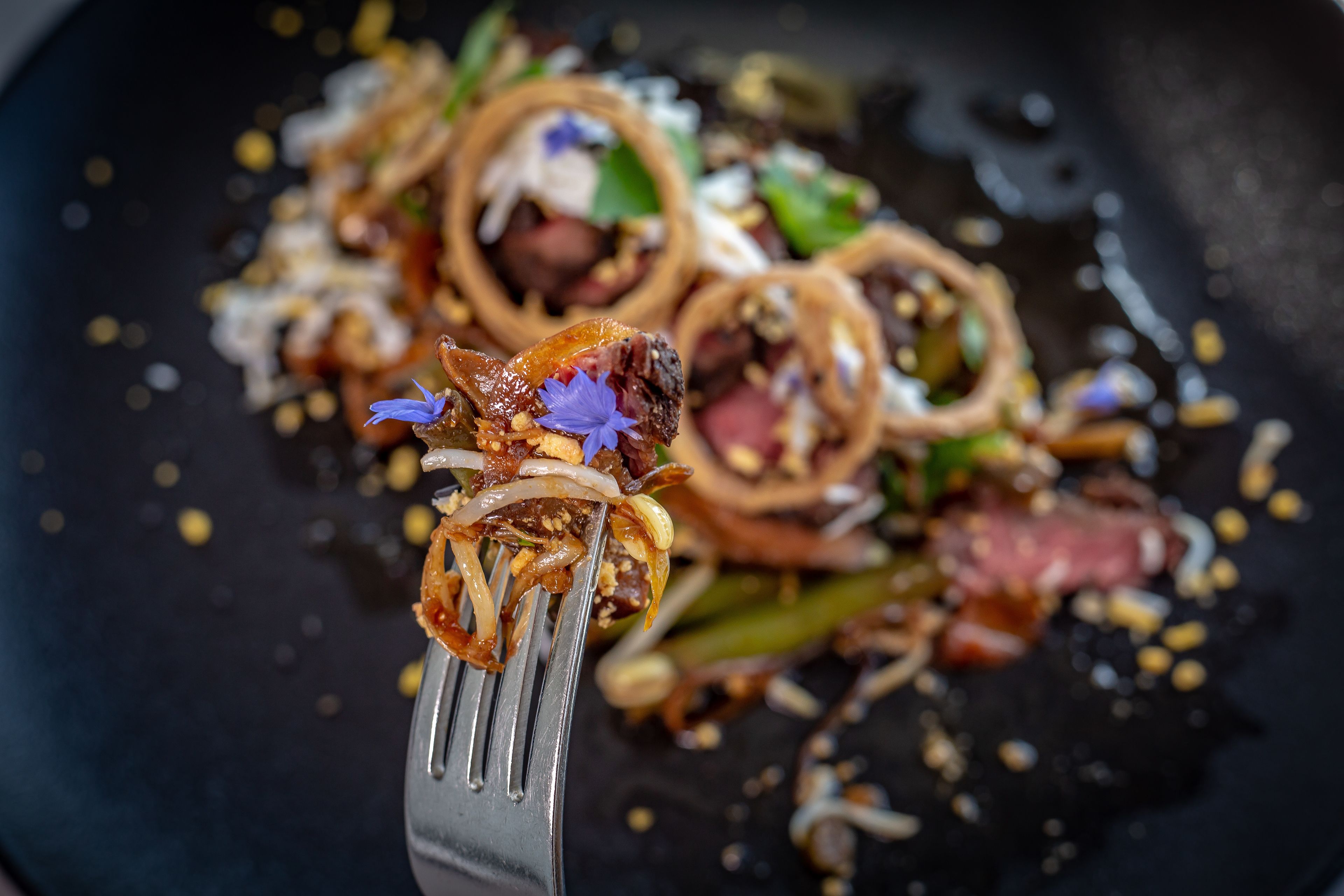 A close-up of a forkful of food with onions, meat, and a flower garnish.