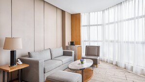 Una habitación de hotel moderna con sofá gris, una mesita redonda pequeña, una mesita de noche con lámpara, un revestimiento de pared de madera y cortinas translúcidas.