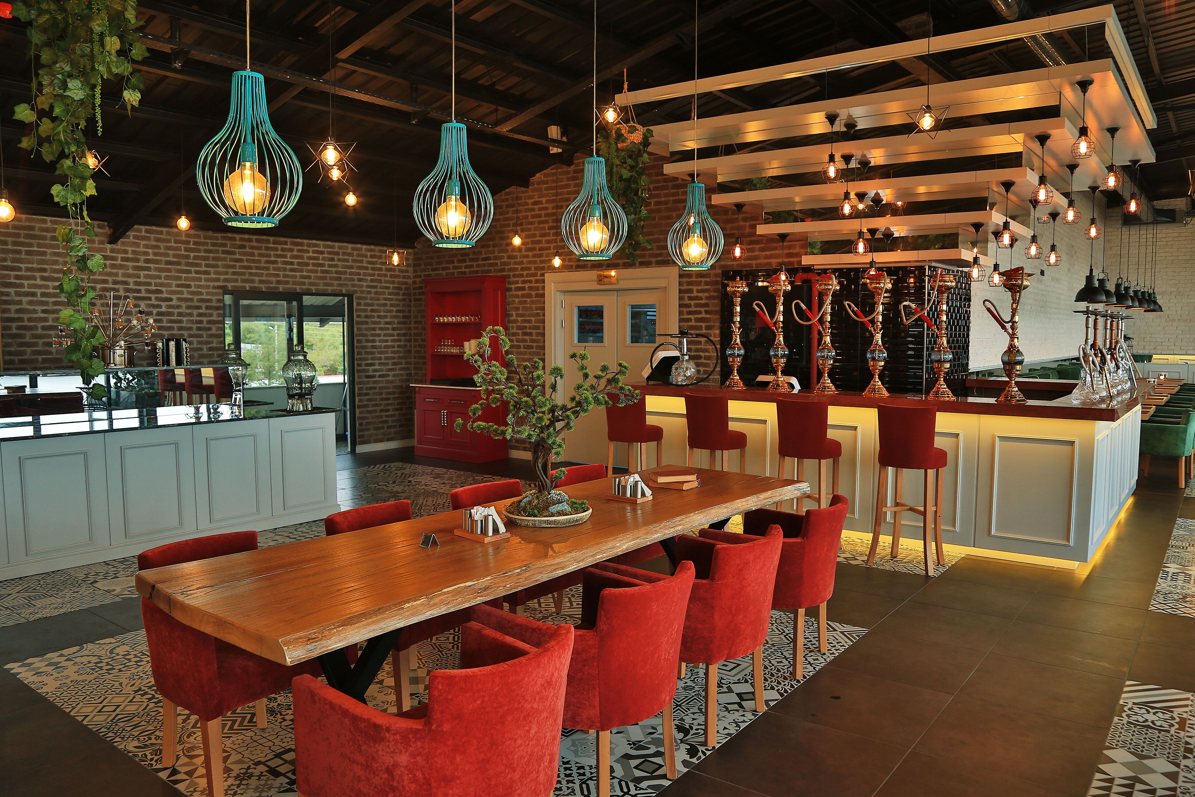 A modern restaurant with a wooden long table, red chairs, hanging green light fixtures, and a bar area with stools.