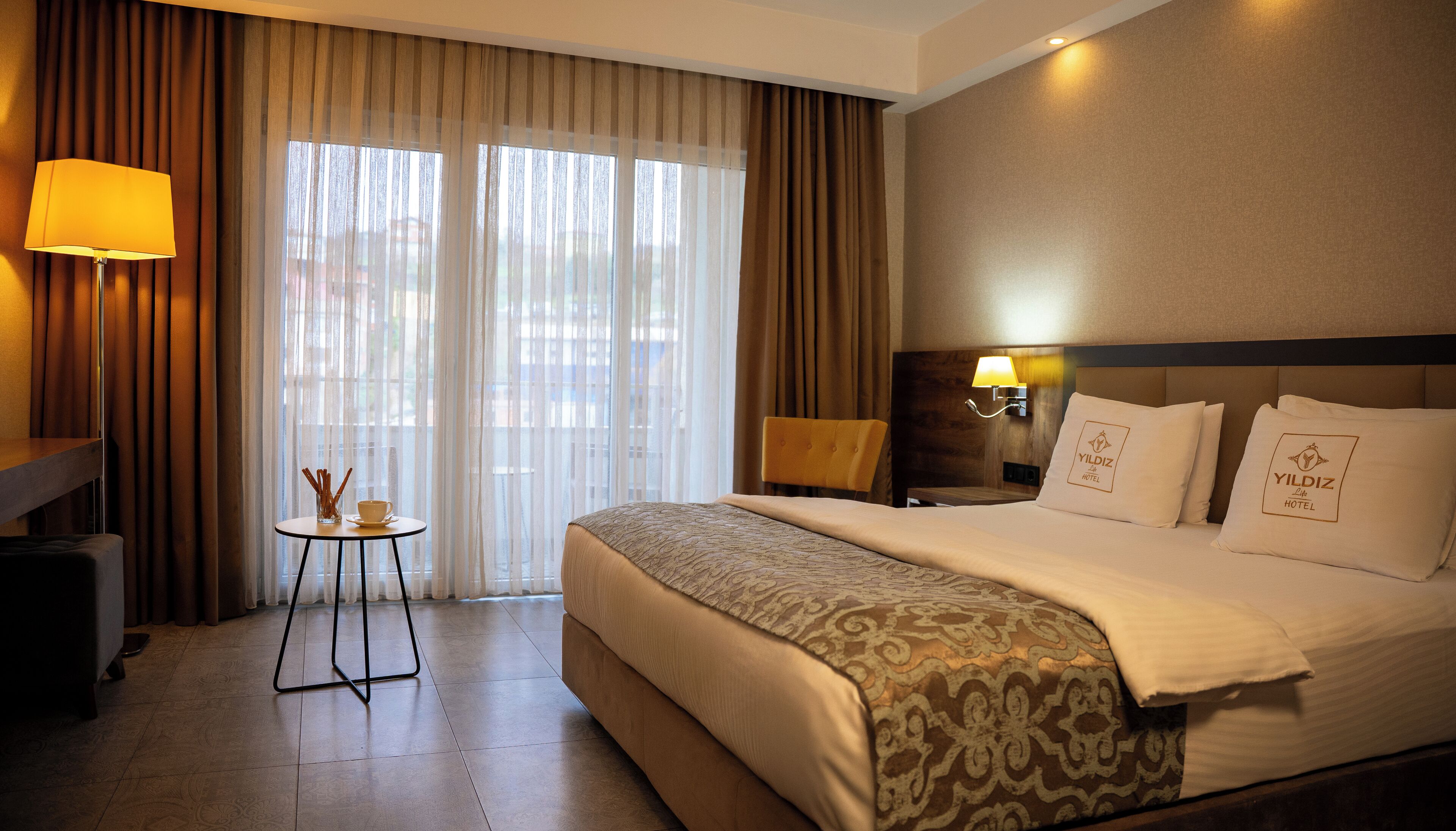 Habitación de hotel con una cama grande, una mesita de noche con lámpara, una pequeña mesa con una taza y un florero, y vistas a través de la ventana.