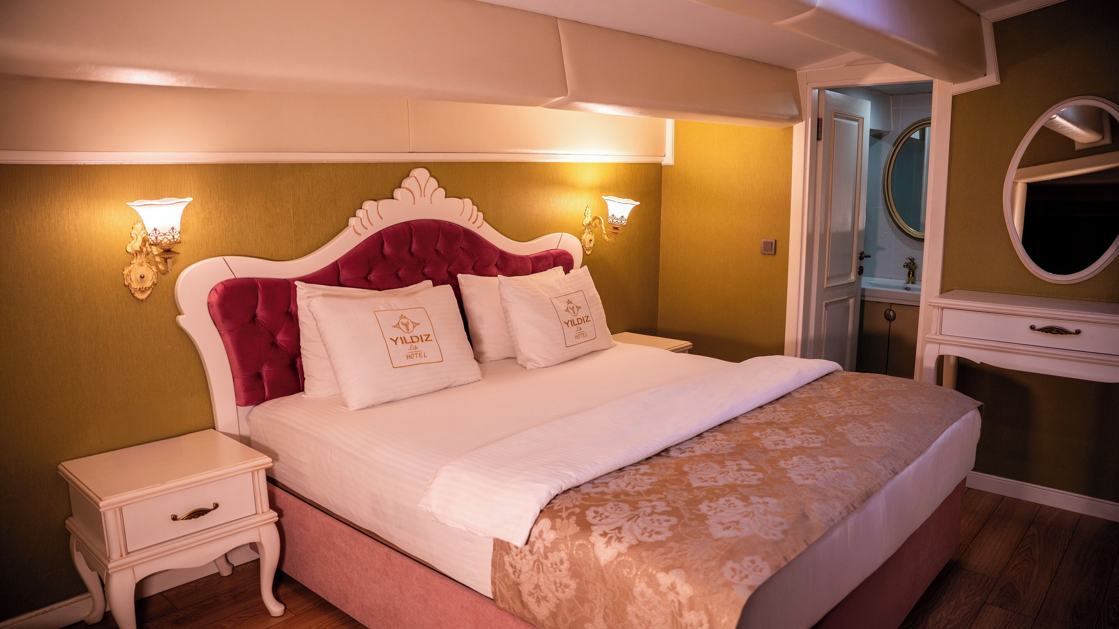A neatly made bed with a red headboard and white bedding, a bedside table, and a wall-mounted mirror.