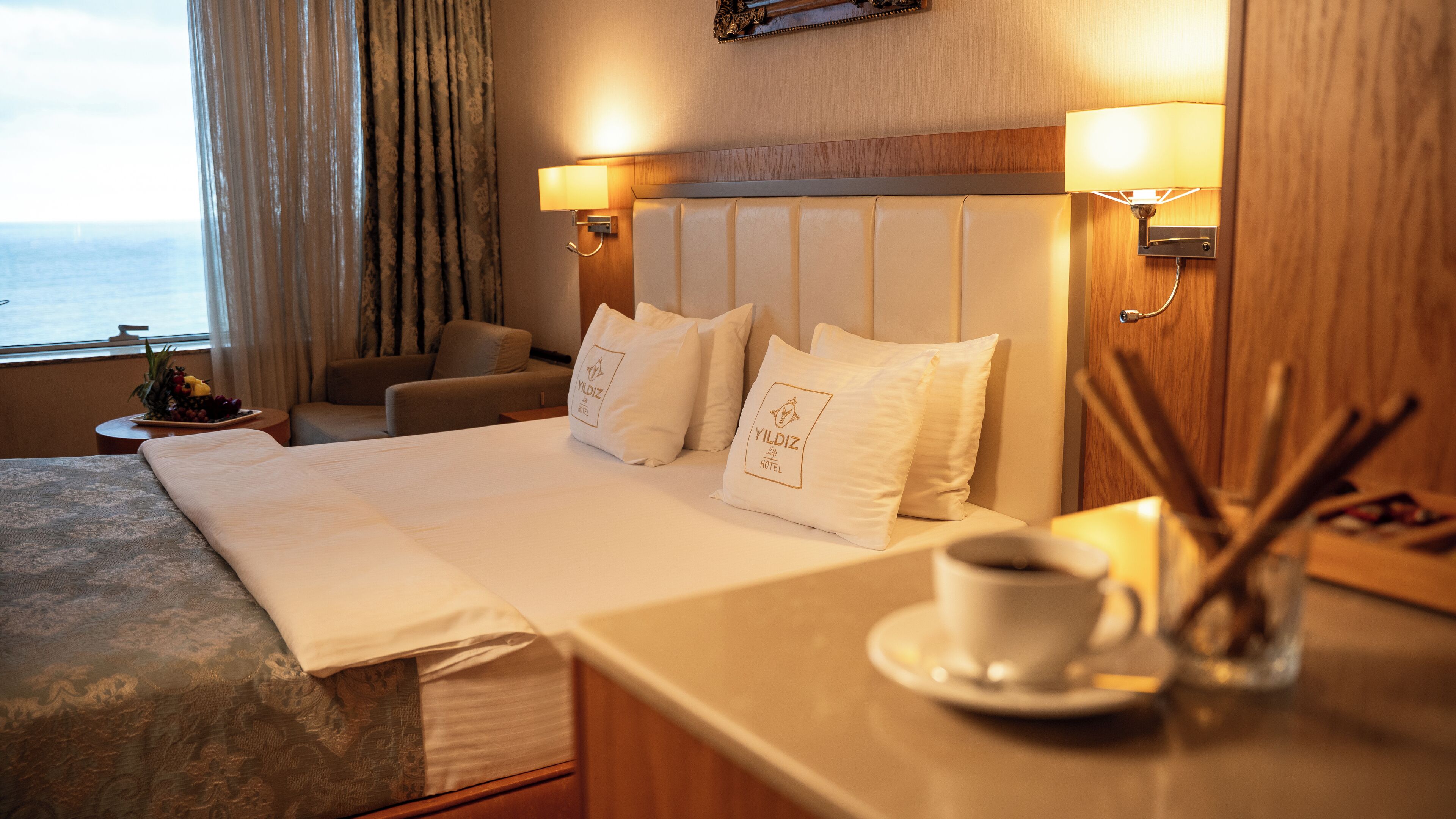 A hotel room with a bed, two bedside lamps, a coffee table with a cup and saucer, and a view of the sea through the window.