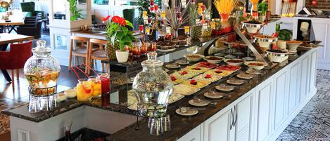 A buffet with a variety of food items and drinks, including fruits, cheeses, and pasta.