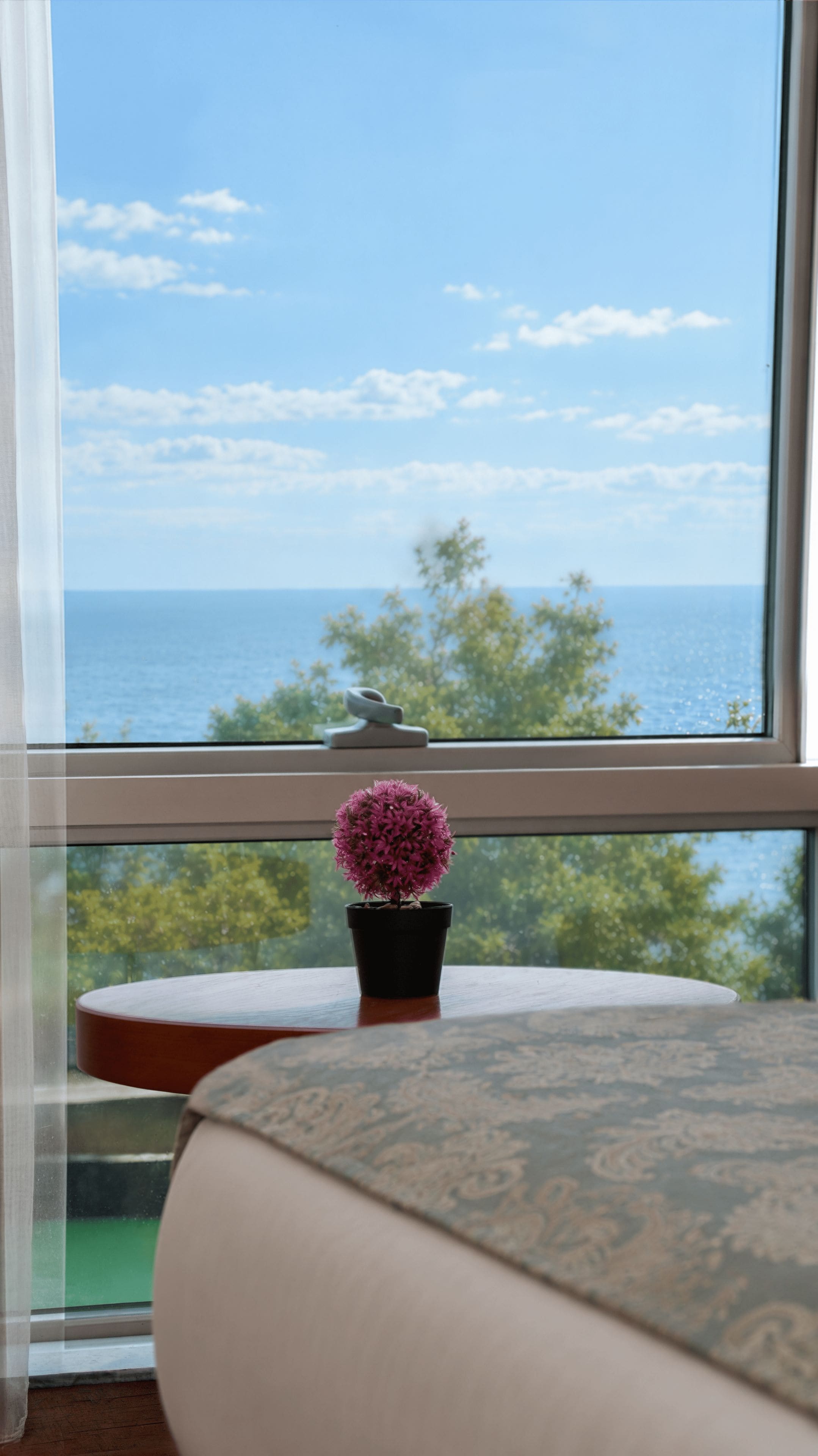 A room with a view of the sea through a large window, a small round table with a potted plant, and a cushion on the floor.