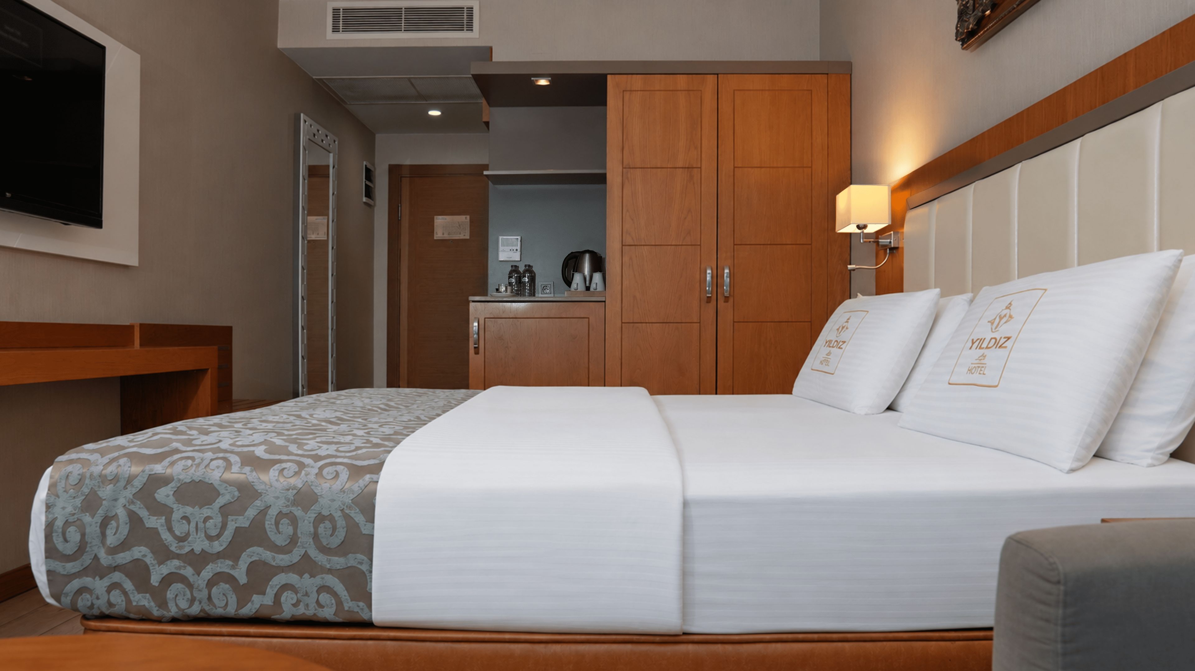 A hotel room with a bed, a desk, a wardrobe, and a kitchenette in the background.