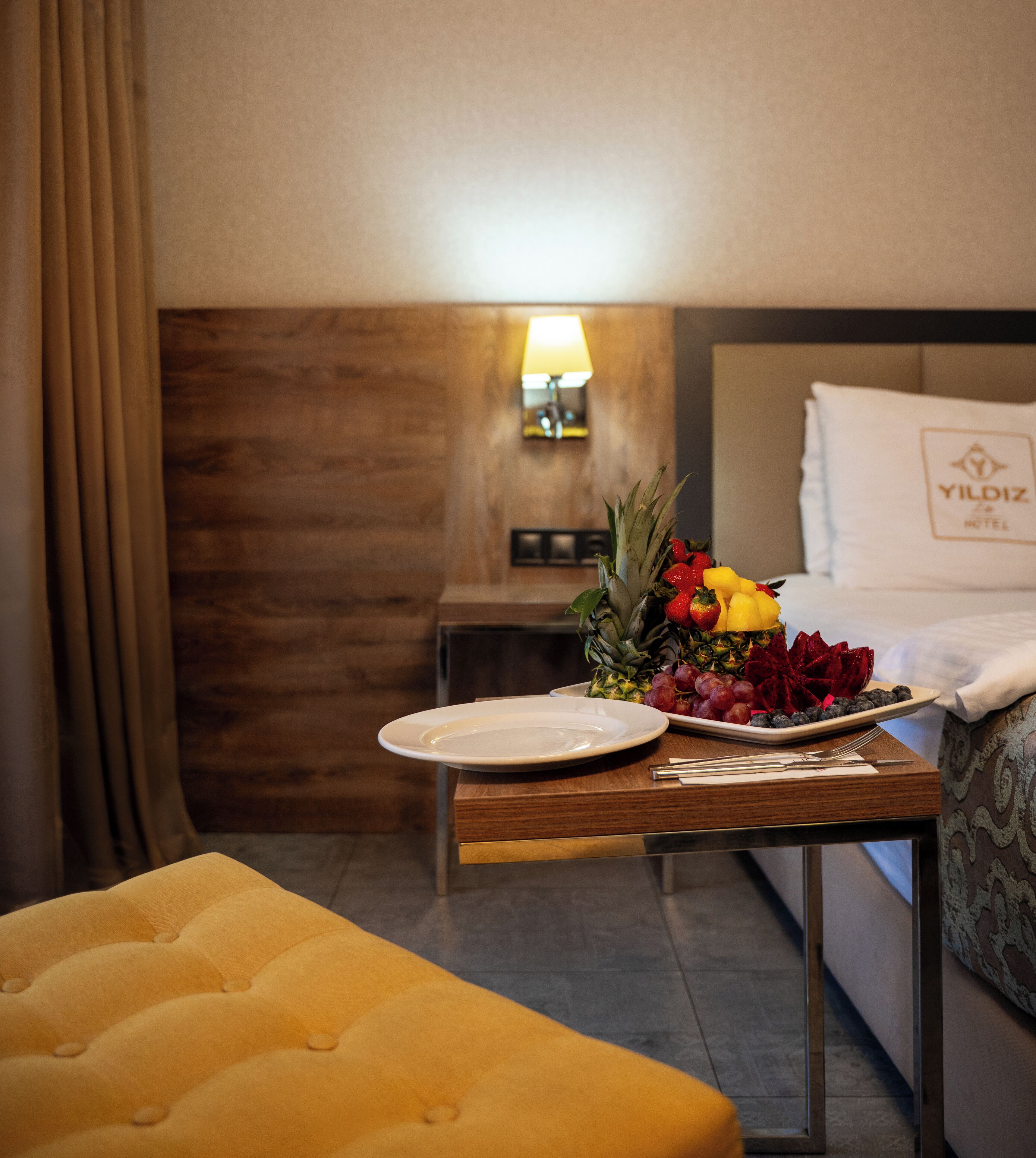 Una habitación de hotel con una cama, una mesita de noche con una bandeja de frutas, una lámpara y un reposapiés amarillo.