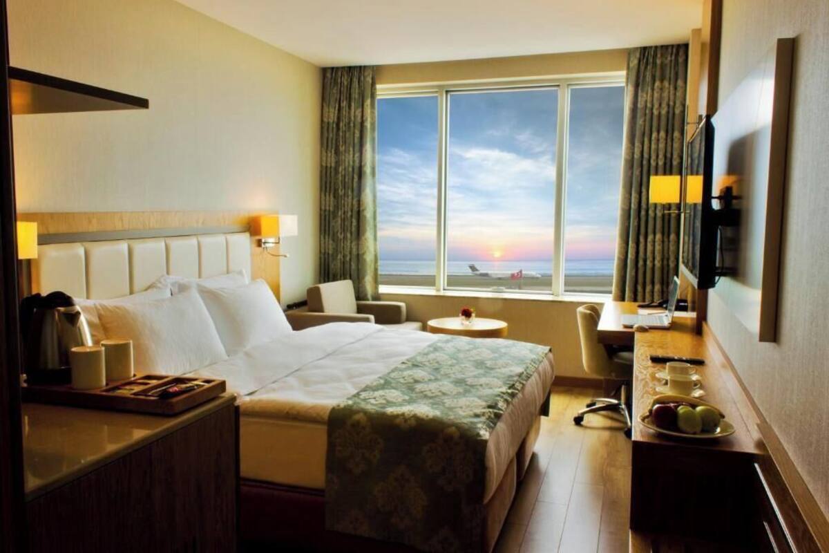 A hotel room with a bed, a desk with a chair, a small table, and a view of the sunset through a large window.