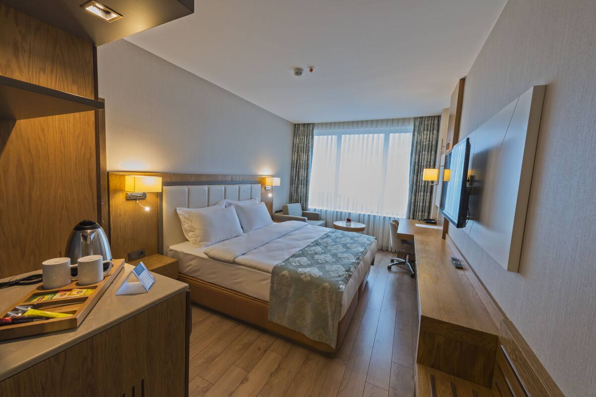 A hotel room with a bed, bedside table, lamp, TV, and a desk with a chair.