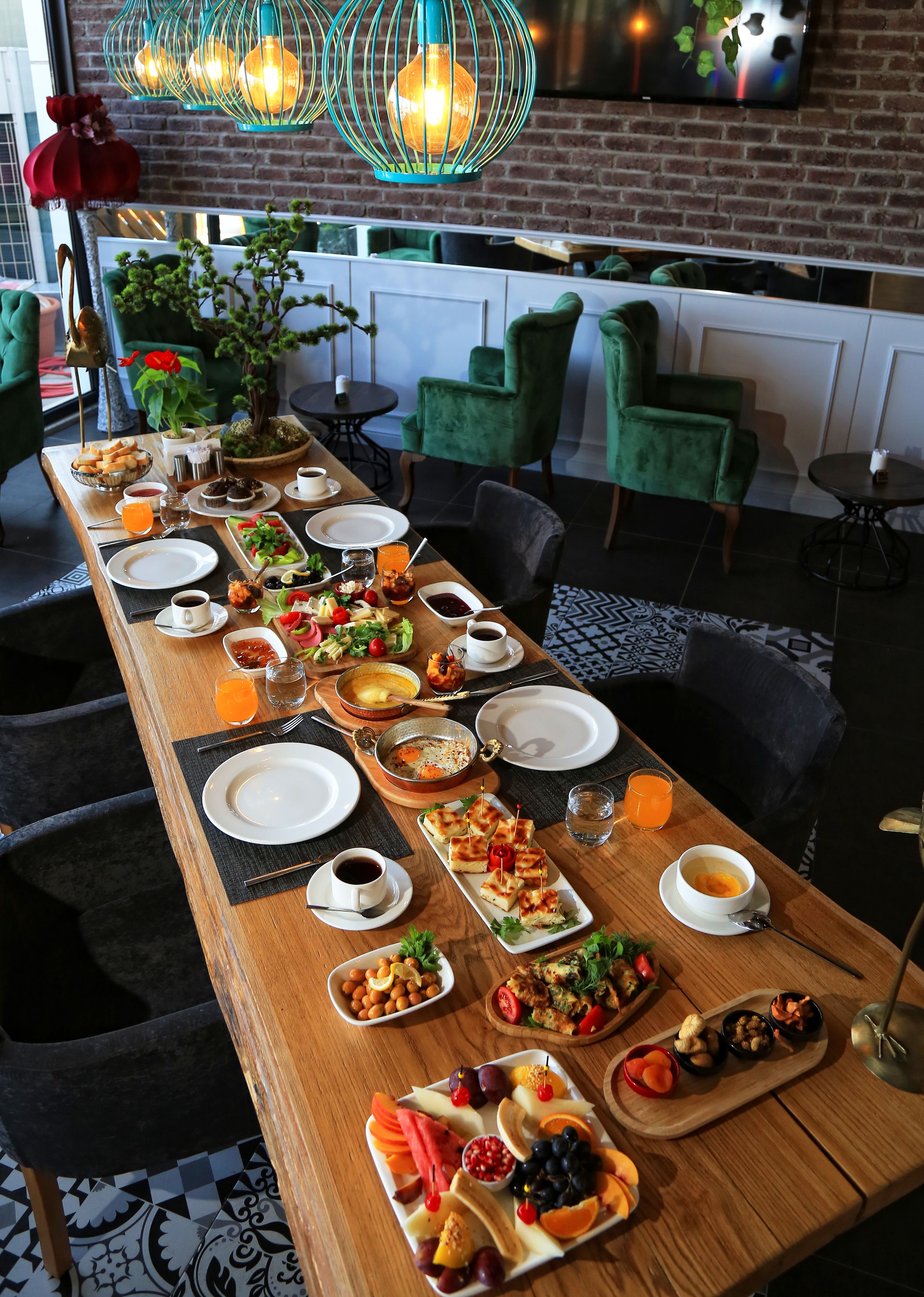 A long dining table set with various breakfast items, surrounded by green chairs in a modern interior with brick walls and a hanging light fixture.