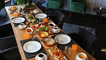 A long dining table set with various breakfast items, surrounded by green chairs in a modern interior with brick walls and a hanging light fixture.