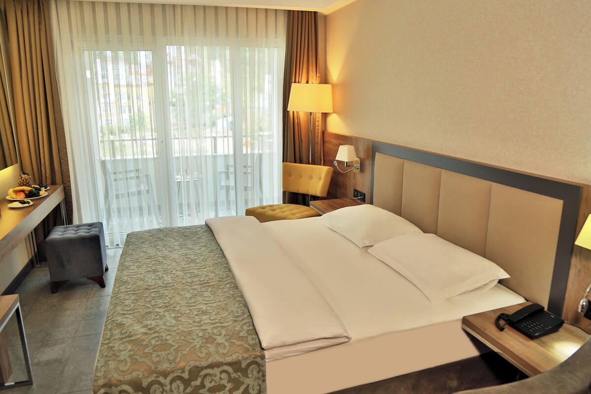 A hotel room with a large bed, a desk with a lamp, a chair, and a small ottoman.