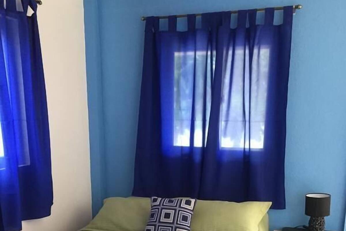 A single bed with a green bedspread and a patterned pillow, a nightstand with a lamp, and blue curtains on a window.