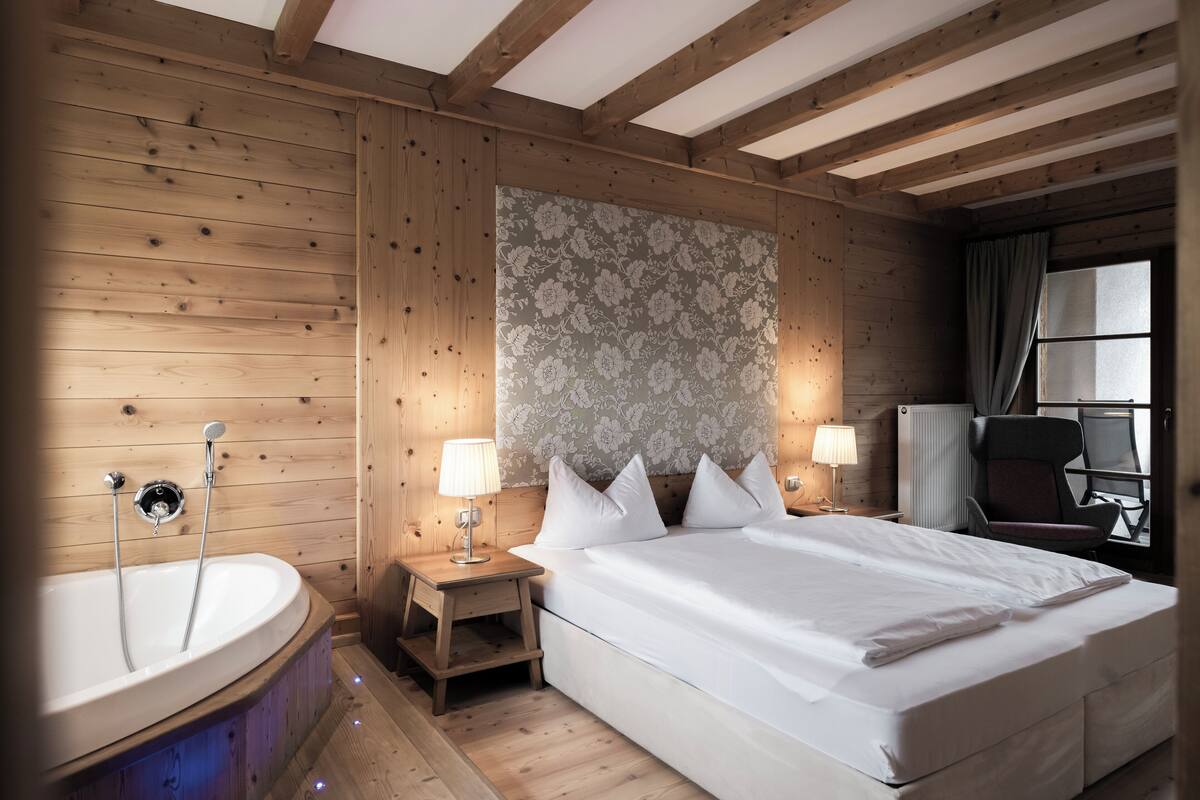 Een kamer met houten lambrisering, een groot bed, een houten nachtkastje, een wandlamp en een raam met gordijnen.