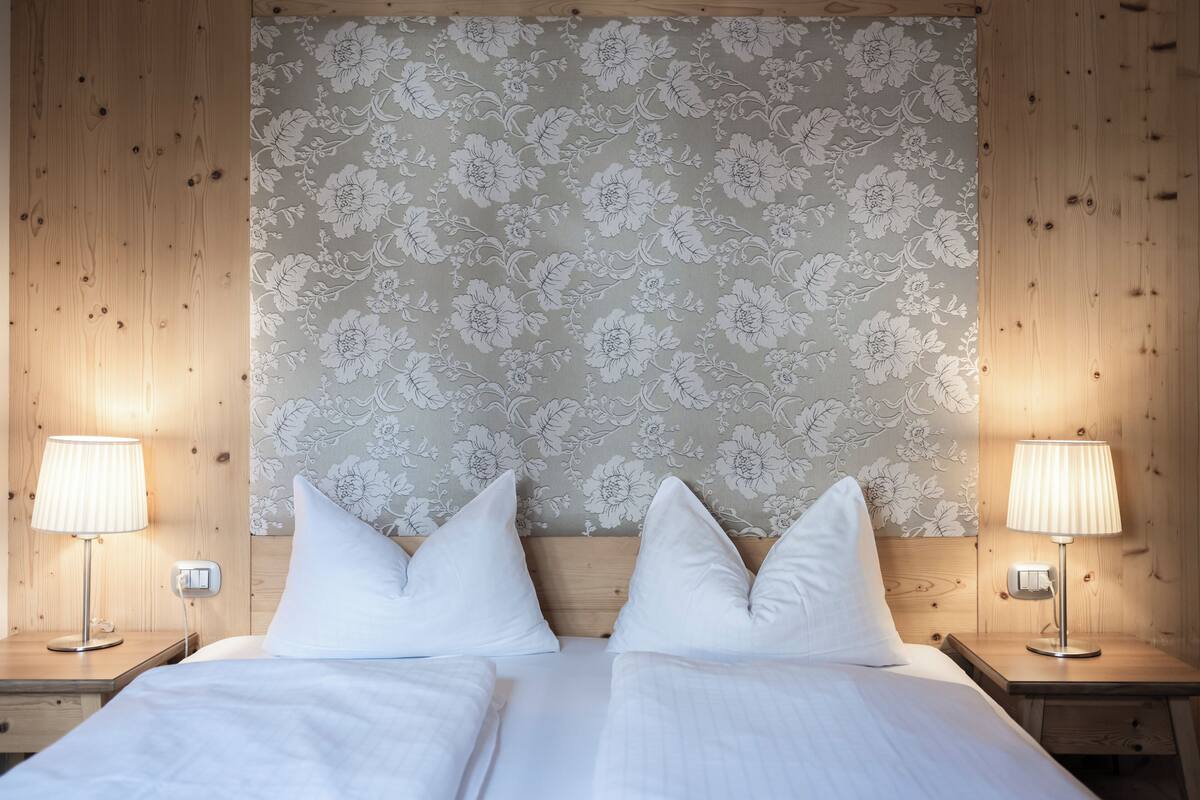Een tweepersoonsbed met witte lakens en twee kussens, met aan weerszijden houten nachtkastjes met lampen, voor een behang met bloemen.