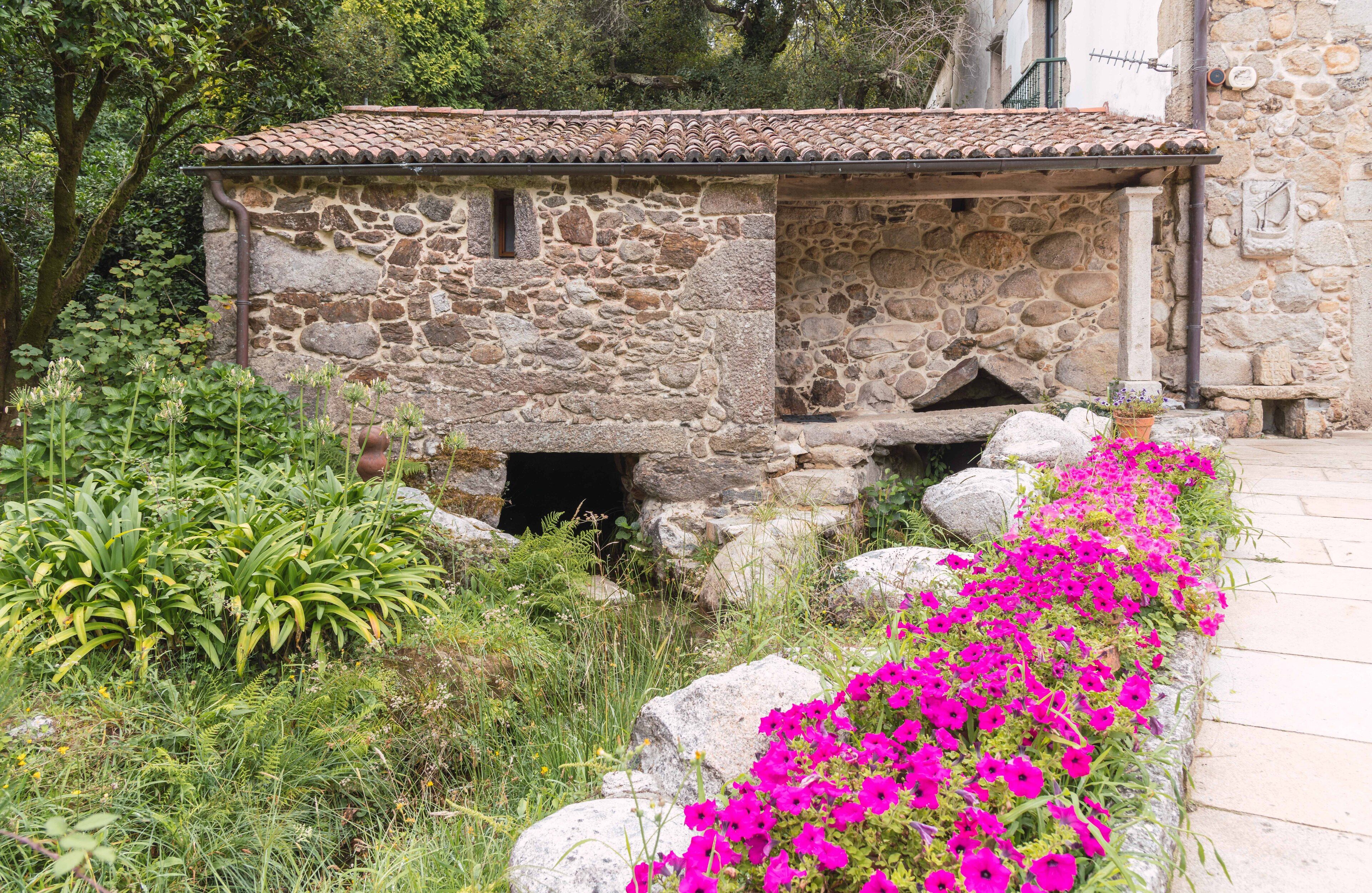 Un edificio de piedra con tejado de baldosas, rodeado de vegetación exuberante y flores coloridas.