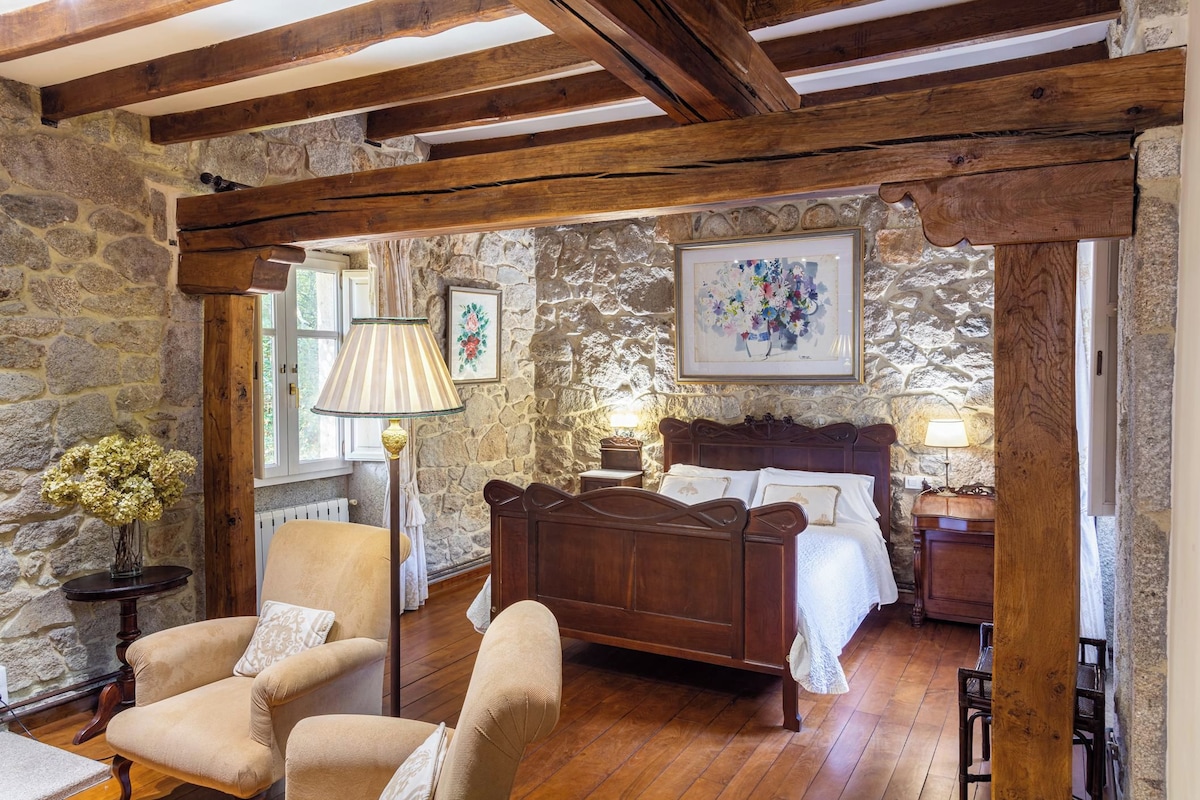 A bedroom with a stone wall, wooden beams, a bed, a chair, and a lamp.