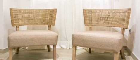 Two wicker chairs with fabric seats and wooden legs, positioned in front of a white curtain.