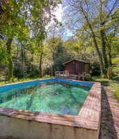 Uma piscina cristalina com um deck de madeira, rodeada por vegetação exuberante e um chalé de madeira.