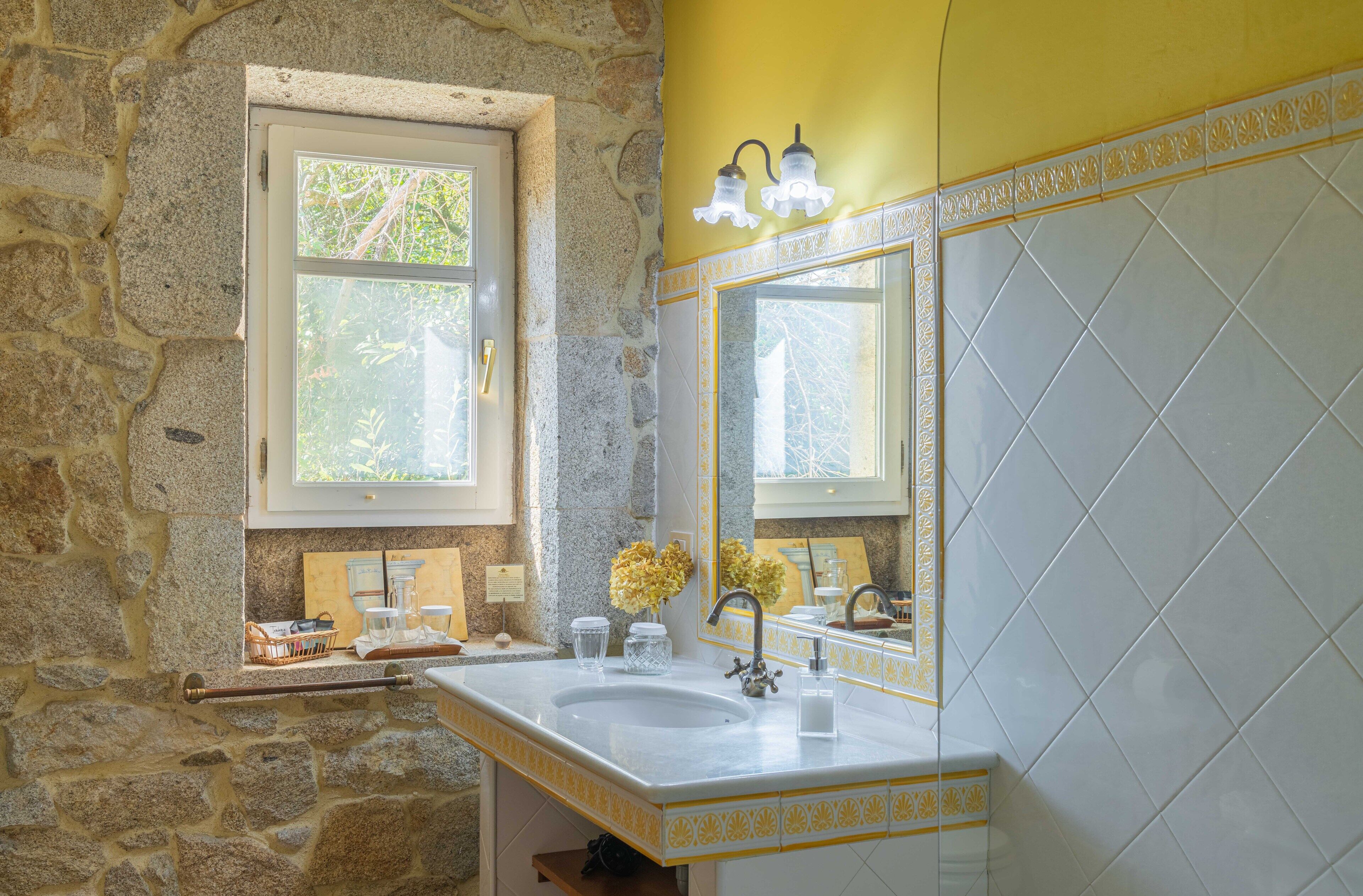 Un baño con pared de piedra, ventana, lavamanos con espejo y azulejos decorativos amarillos.