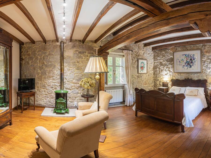 A room with a stone wall, wooden beams, a bed, a chair, a lamp, and a television.