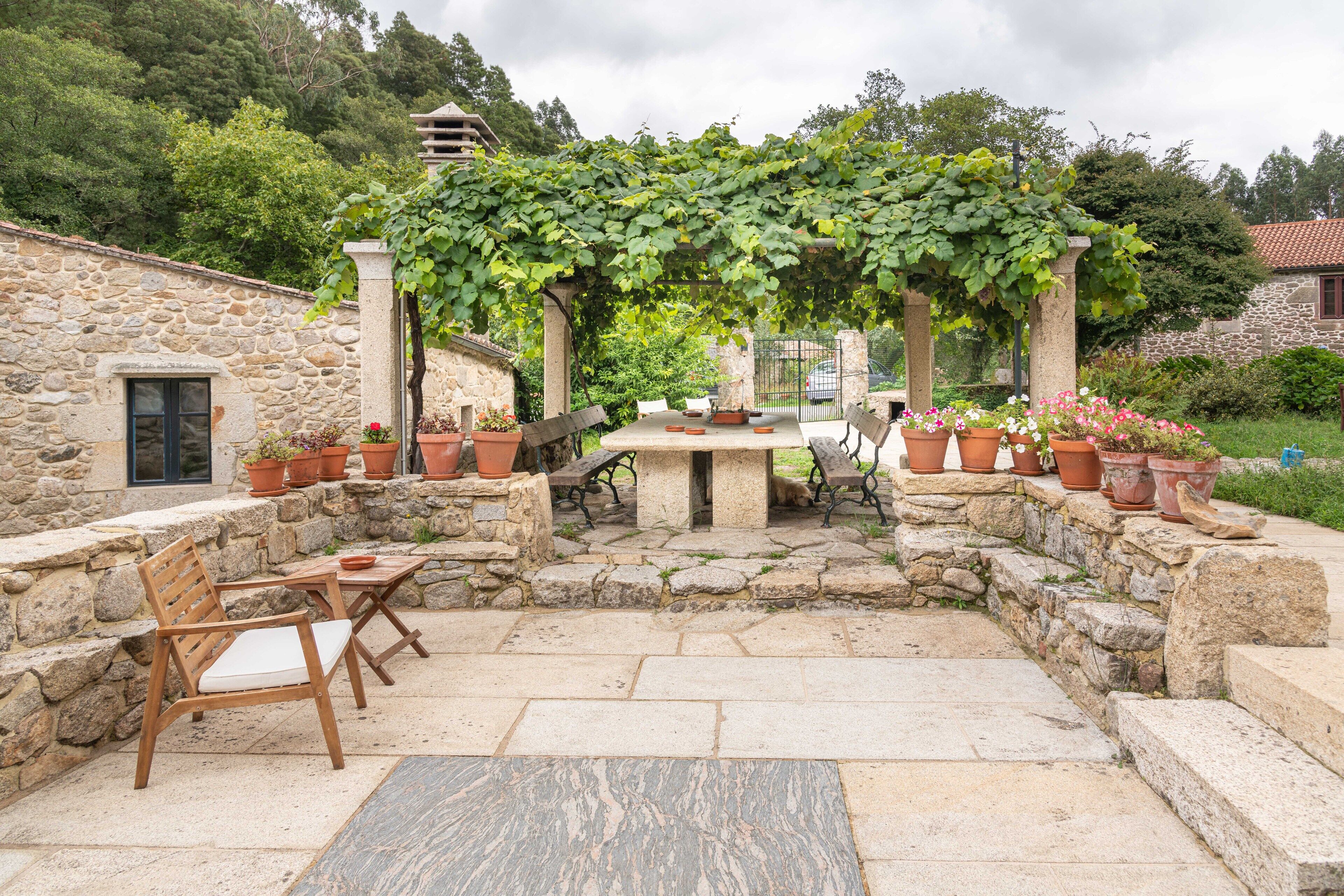 A stone patio with a table and chairs, surrounded by potted plants and a pergola with vines.