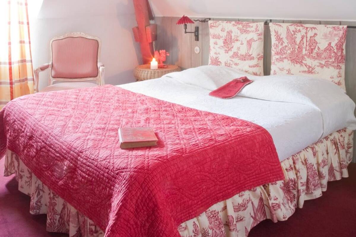 A bedroom with a sloped ceiling, a bed with a red quilt, a chair, and a nightstand with a candle.