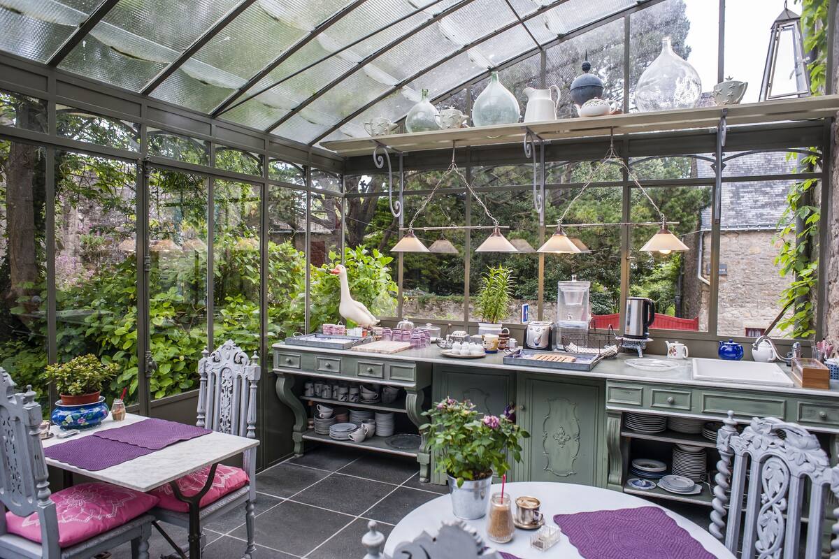A conservatory with a glass roof, green cabinets, and a variety of plants and kitchenware.
