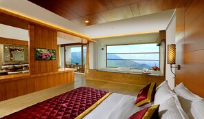 A modern hotel room with a large bed, a wooden desk, a large window with a mountain view, and a balcony with a view of the sea. - PLUM JUDY - THE LEISURE HOTEL (Devikolam)