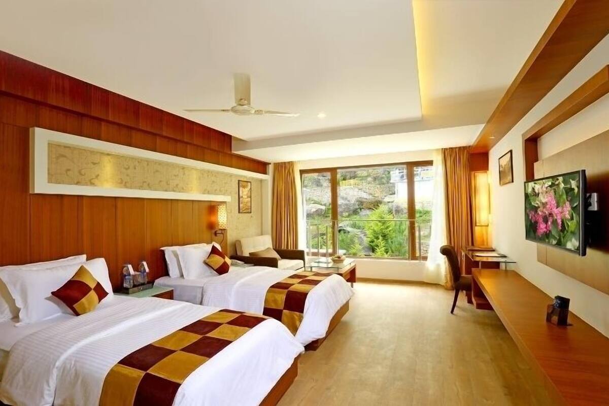 A hotel room with two beds, a desk, a chair, a TV, and a large window offering a view of greenery.