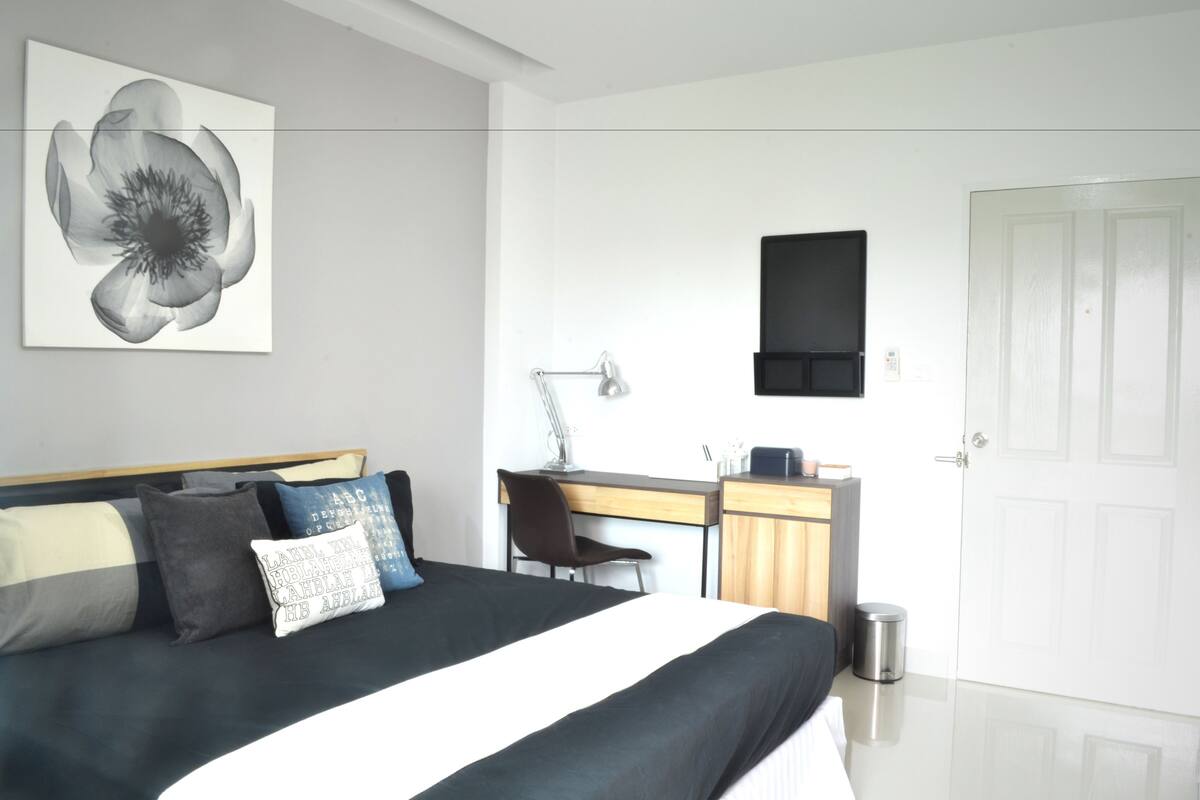 A modern hotel room with a large bed, a desk with a chair, a small trash bin, and a door.