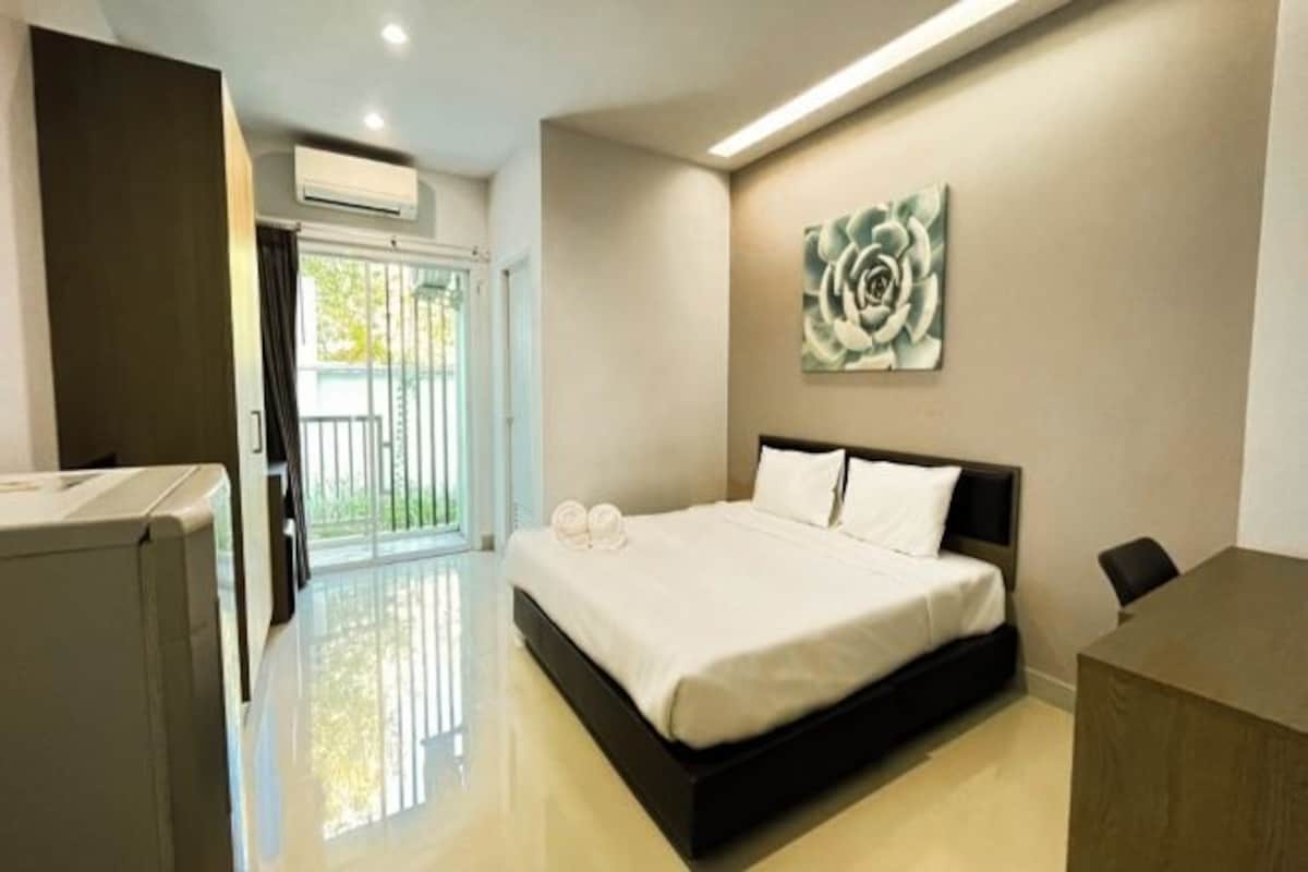 A modern hotel room with a large bed, a desk, and a sliding glass door leading to a balcony.