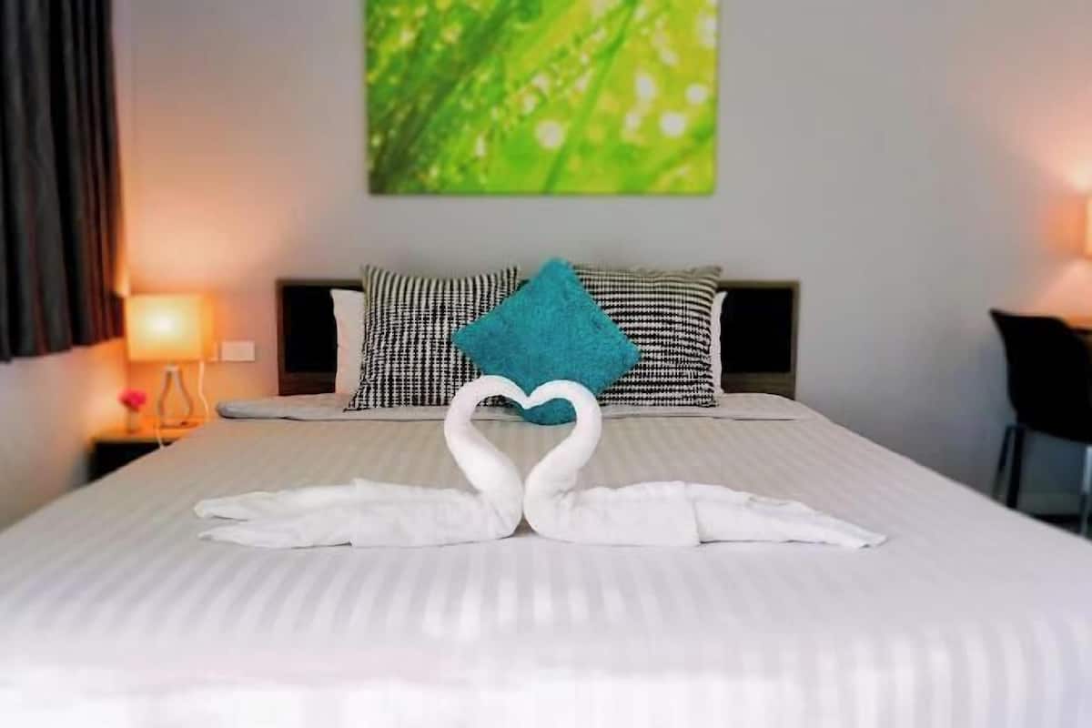 A neatly made bed with a heart-shaped towel arrangement, a green pillow, and a wall painting.