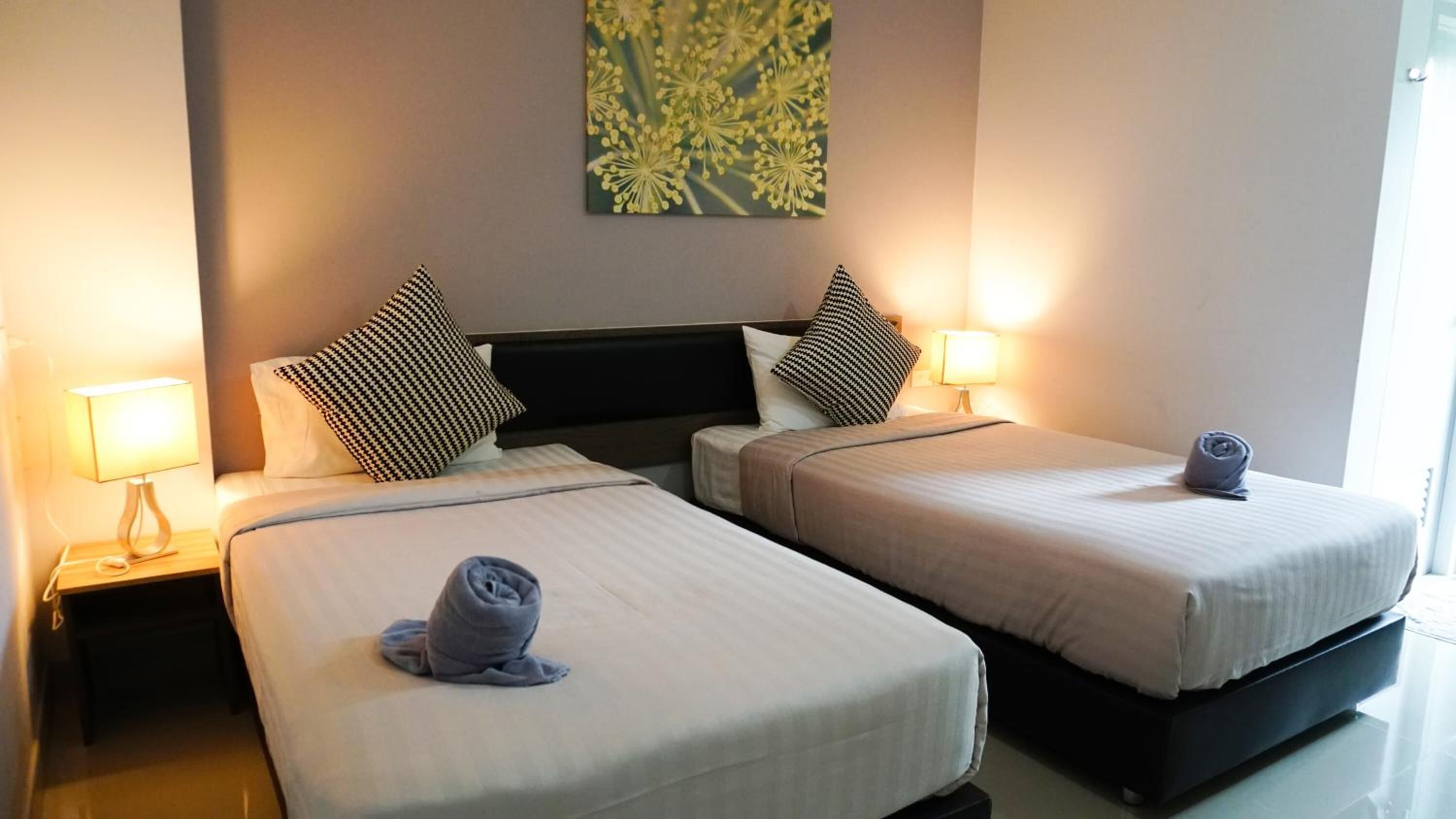 A hotel room with two single beds, each with a pillow and a rolled towel. There is a nightstand with a lamp on each side of the beds.