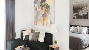 Ein modernes Wohnzimmer mit einer Couch, einem kleinen Tisch und einer Lampe. Im Hintergrund ist ein Bett mit einer grauen Tagesdecke zu sehen.