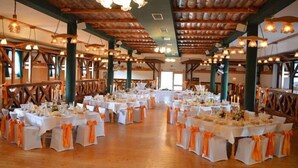 Un salón de banquetes con varias mesas redondas cubiertas con manteles blancos y cintas anaranjadas, vigas de madera en el techo y ventanas grandes.