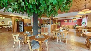 Un restaurante con mesas y sillas de madera, un gran árbol de hojas verdes y un piso de baldosas.