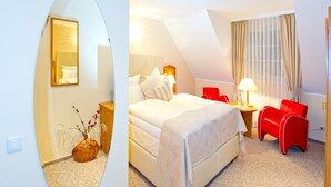 Una habitación de hotel con una cama, un sillón rojo, una mesita de noche con una lámpara y un espejo en la pared.