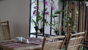 Un comedor con sillas de bambú y una mesa dispuesta con un posavasos y una servilleta. Hay orquídeas en macetas sobre un soporte de metal negro detrás de la mesa.