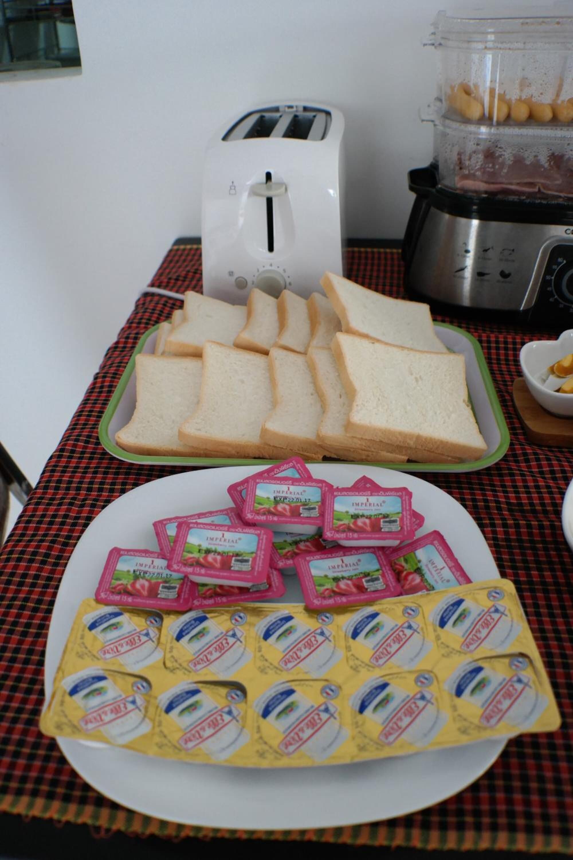 A table with a checkered tablecloth, a toaster, a blender, and plates of sliced bread and cheese packets.