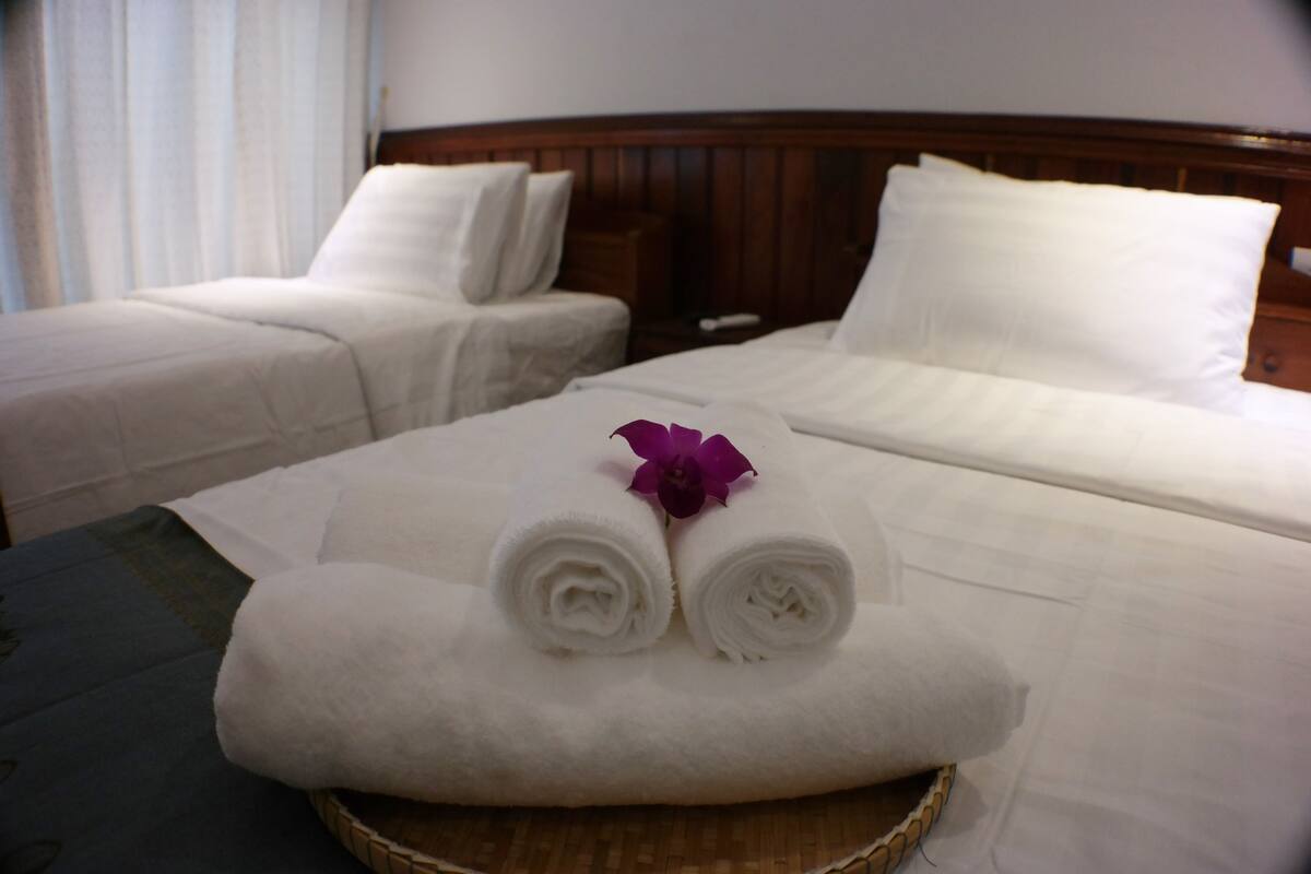 A hotel room with two single beds, each with white linens and pillows, and a large bed with rolled white towels and a purple flower.