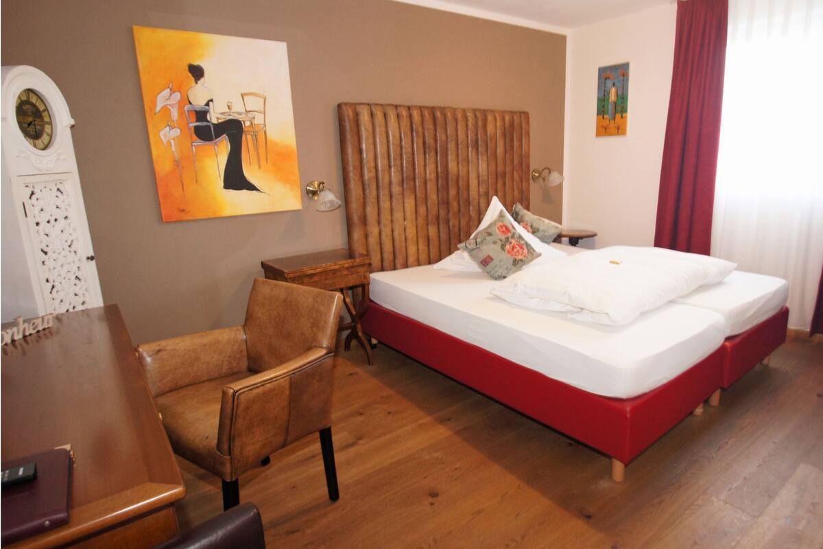 Habitación de hotel con piso de madera, cama con ropa de cama roja, cabecera de madera, escritorio con silla y un cuadro en la pared.