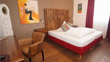A hotel room with a wooden floor, a bed with red bedding, a wooden headboard, a desk with a chair, and a painting on the wall.