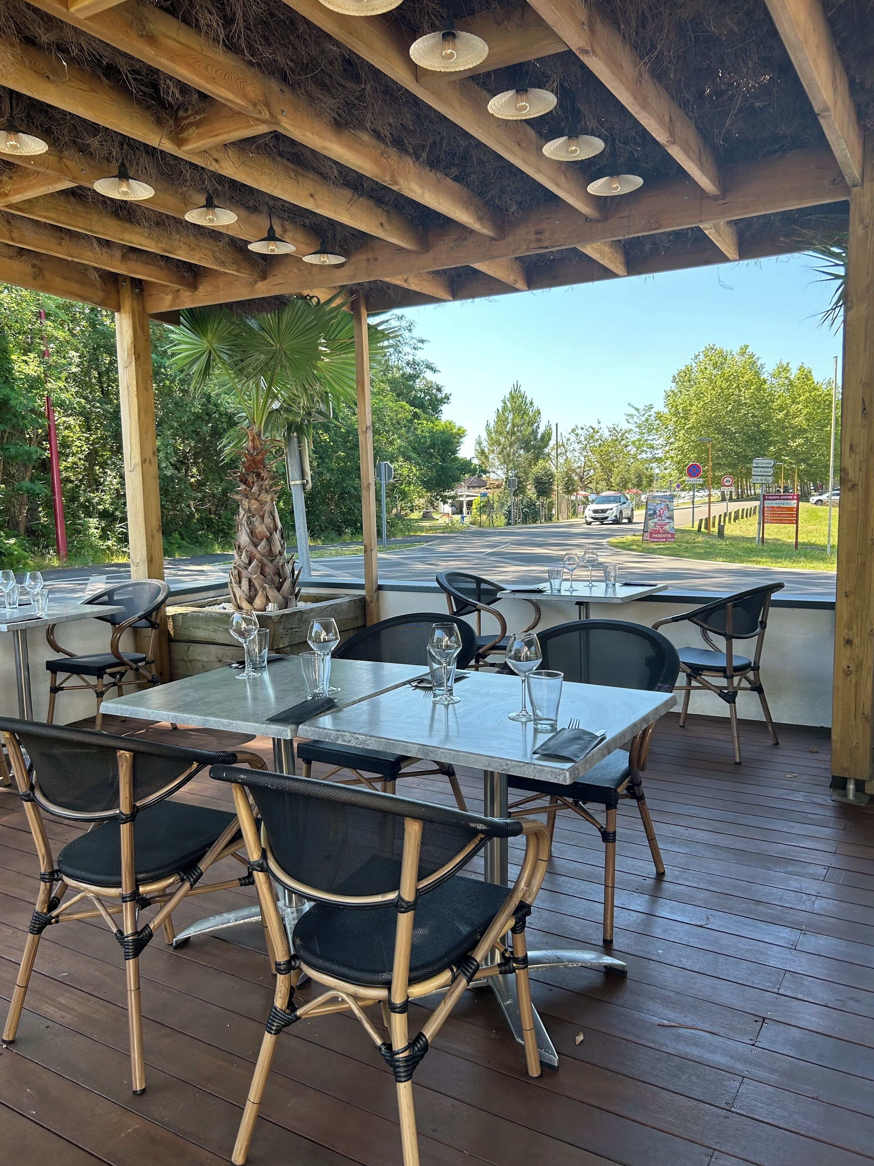 Un espace de restauration en plein air avec des tables et des chaises, un plafond en bois et une vue sur la rue et la verdure.
