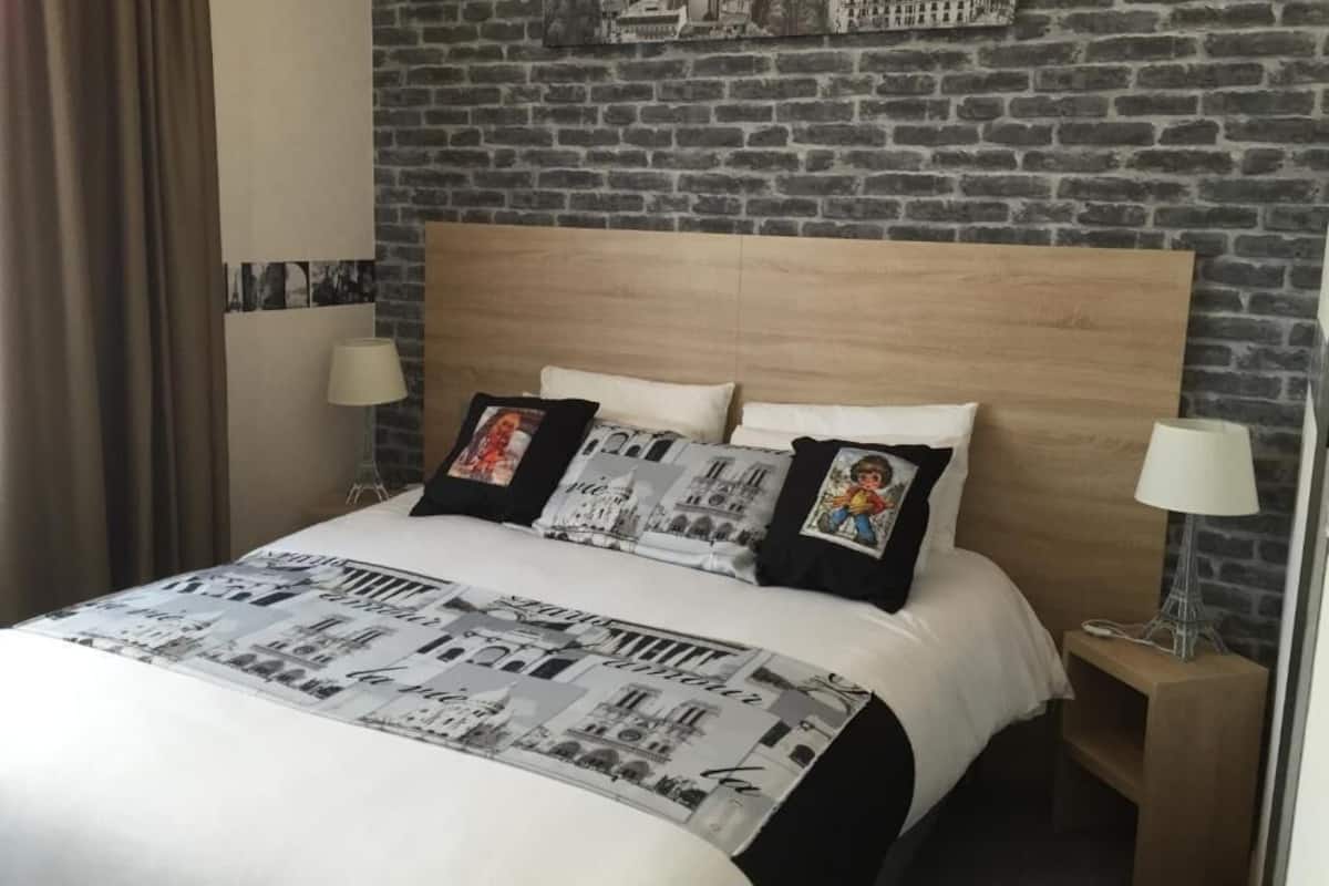 A hotel room with a brick wall, a wooden headboard, a bed with a cityscape bedspread, two bedside lamps, and a nightstand with a lamp.