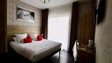 A hotel room with a large bed, a wooden headboard, a mounted picture, a bedside table with a lamp, and a window with curtains.