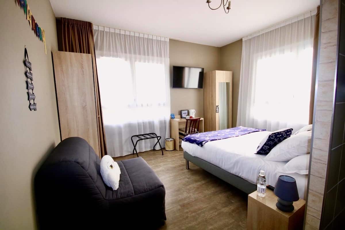 A hotel room with a bed, a desk, a chair, a TV, and a small table with a lamp and a bottle.