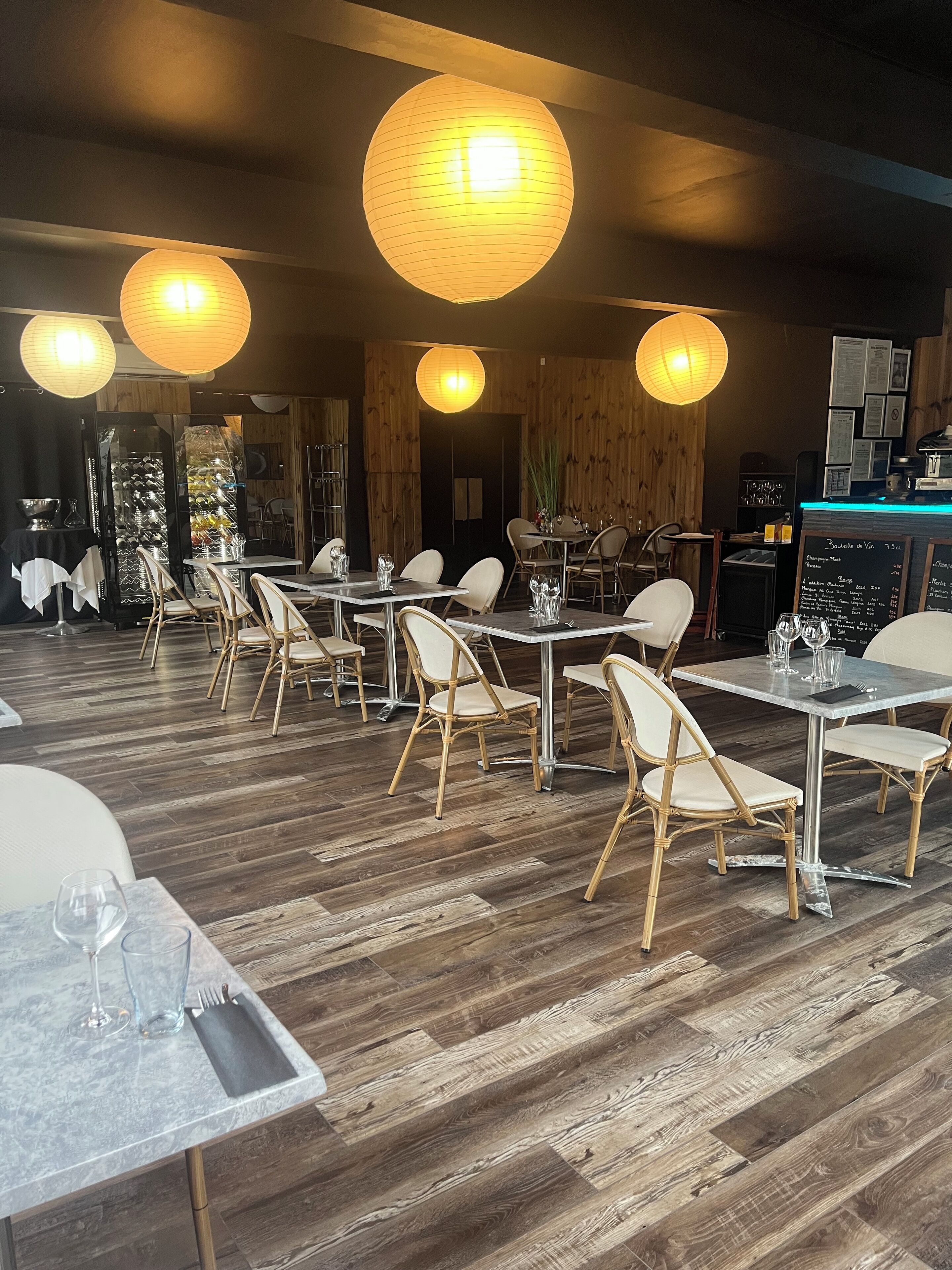 A restaurant interior with wooden flooring, white tables, and chairs, and hanging paper lanterns.